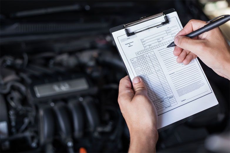 Mechanic filling out a vehicle inspection checklist on a clipboard in front of a car engine.